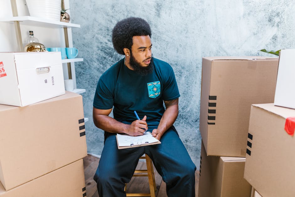 A man with a dark beard and long black hair tied back, wearing a grey t-shirt and beige trousers, sits on the floor inside a property surrounded by moving boxes with red and yellow markings, indicativ