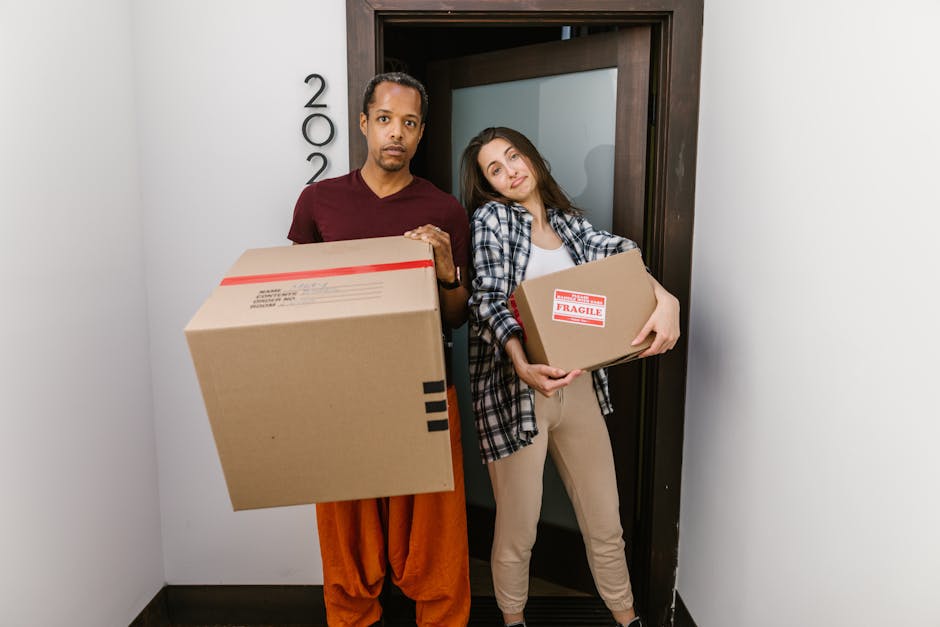A man and a woman stand inside a home near a doorway, each holding a cardboard box used for packing during a house removal. The man, dressed in an orange pair of trousers and a maroon shirt, holds a l